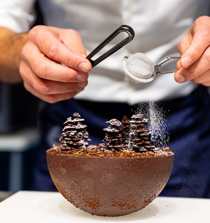 Zoom in on a pastry depicting a forest of fir trees sprinkled with powdered sugar.