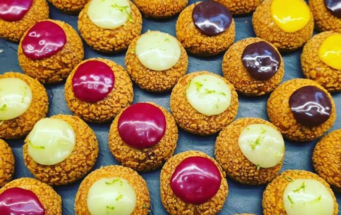 Craquelin choux pastries topped with yellow, white and purple icing, arranged on a baking tray.